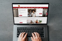 A person is using a MacBook Pro to browse a banking website. The screen displays a login section, promotional offers, and an image of a smiling couple in the top section. The design includes a red color scheme, with navigation links for various banking services.