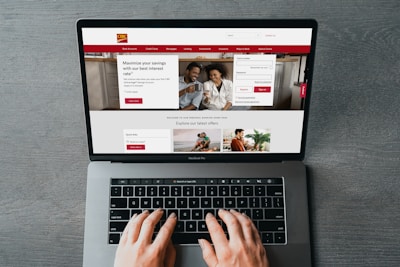 A person is using a MacBook Pro to browse a banking website. The screen displays a login section, promotional offers, and an image of a smiling couple in the top section. The design includes a red color scheme, with navigation links for various banking services.