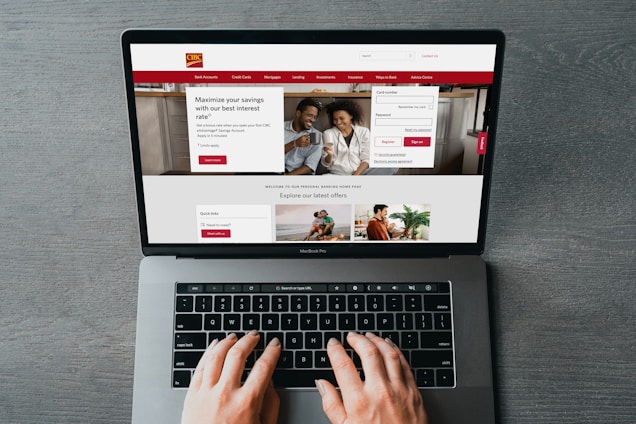 A person is using a MacBook Pro to browse a banking website. The screen displays a login section, promotional offers, and an image of a smiling couple in the top section. The design includes a red color scheme, with navigation links for various banking services.