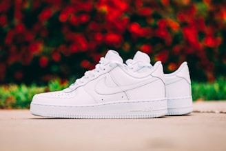 A pair of pristine white sneakers placed on a smooth outdoor surface. The sneakers have a sleek design, with a prominent logo on the side. In the background, out of focus, there is a burst of red and green colors, suggesting foliage or flowers.