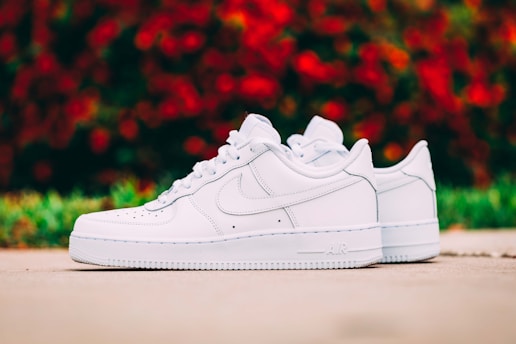 A pair of pristine white sneakers placed on a smooth outdoor surface. The sneakers have a sleek design, with a prominent logo on the side. In the background, out of focus, there is a burst of red and green colors, suggesting foliage or flowers.