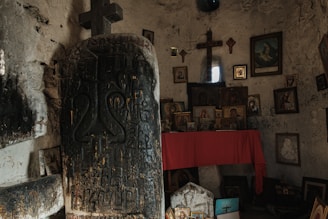A dimly lit room with religious icons and paintings displayed on a wall. A stone slab with engravings and a cross stands prominently in the foreground. A red cloth covers a table near the wall, adding a touch of color to the otherwise muted tones of the space.