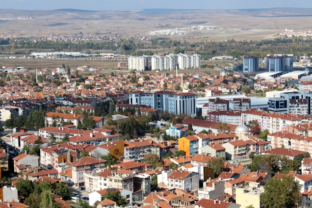 A sprawling urban landscape with a mix of residential buildings and mid-rise apartments featuring red-tiled roofs. The city stretches across a flat plane, flanked by a distant view of rolling hills. A few taller, modern buildings rise amidst older, more traditional structures. Vegetation is interspersed throughout the area, with trees lining some of the streets.
