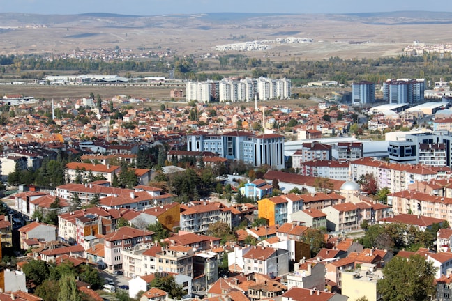 A sprawling urban landscape with a mix of residential buildings and mid-rise apartments featuring red-tiled roofs. The city stretches across a flat plane, flanked by a distant view of rolling hills. A few taller, modern buildings rise amidst older, more traditional structures. Vegetation is interspersed throughout the area, with trees lining some of the streets.