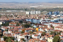A sprawling urban landscape with a mix of residential buildings and mid-rise apartments featuring red-tiled roofs. The city stretches across a flat plane, flanked by a distant view of rolling hills. A few taller, modern buildings rise amidst older, more traditional structures. Vegetation is interspersed throughout the area, with trees lining some of the streets.