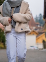 A person dressed warmly in a beige, textured coat and a large grey scarf, paired with white pants. The background consists of a blurred outdoor scene with autumnal tones, featuring muted orange roofs and green foliage.