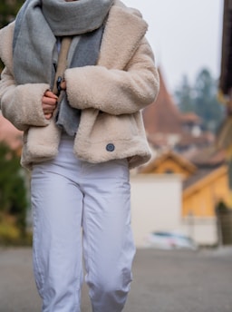 A person dressed warmly in a beige, textured coat and a large grey scarf, paired with white pants. The background consists of a blurred outdoor scene with autumnal tones, featuring muted orange roofs and green foliage.