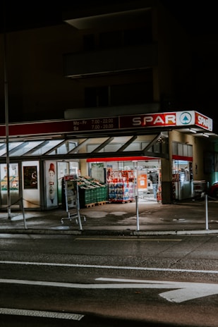 A small supermarket is illuminated at night with light shining from the store’s interior. The parking area is empty, and a shopping cart can be seen outside. The supermarket has a red sign with white lettering.