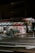 A small supermarket is illuminated at night with light shining from the store’s interior. The parking area is empty, and a shopping cart can be seen outside. The supermarket has a red sign with white lettering.