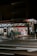 A small supermarket is illuminated at night with light shining from the store’s interior. The parking area is empty, and a shopping cart can be seen outside. The supermarket has a red sign with white lettering.