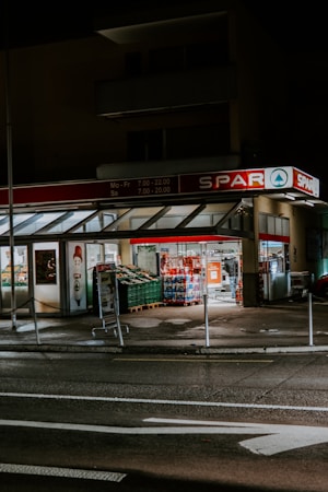 A small supermarket is illuminated at night with light shining from the store’s interior. The parking area is empty, and a shopping cart can be seen outside. The supermarket has a red sign with white lettering.