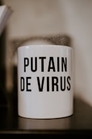 A white mug with bold black text displaying an expression in French that conveys frustration or annoyance related to a virus. The mug is placed on a dark surface with a neutral blurry background.