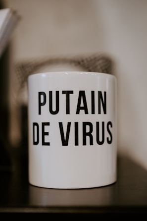 A white mug with bold black text displaying an expression in French that conveys frustration or annoyance related to a virus. The mug is placed on a dark surface with a neutral blurry background.