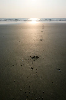 Footprints in soft sand leading toward a distant lighthouse under a blue sky