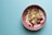 A bowl filled with yogurt topped with a variety of toppings including banana chips, blueberries, raspberries, granola, and nuts is placed on a light teal surface. The bowl is pink and contrasts with the muted background color.