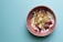A bowl filled with yogurt topped with a variety of toppings including banana chips, blueberries, raspberries, granola, and nuts is placed on a light teal surface. The bowl is pink and contrasts with the muted background color.
