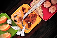 A selection of three colorful trays is filled with a variety of healthy snacks, including orange slices, nuts, seeds, and a measuring tape is artfully draped across them, suggesting themes of dieting or nutrition.