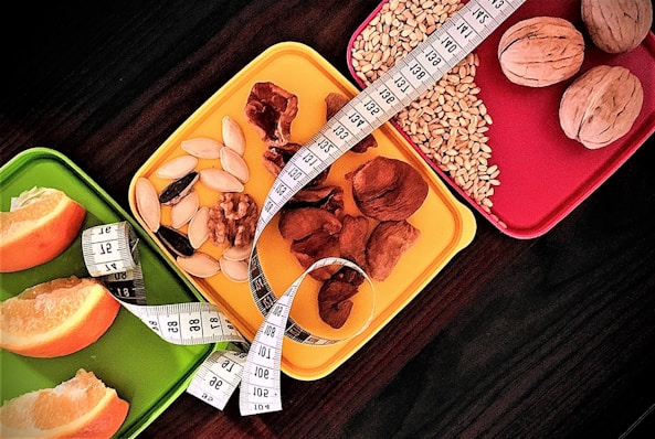 A selection of three colorful trays is filled with a variety of healthy snacks, including orange slices, nuts, seeds, and a measuring tape is artfully draped across them, suggesting themes of dieting or nutrition.