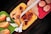 A selection of three colorful trays is filled with a variety of healthy snacks, including orange slices, nuts, seeds, and a measuring tape is artfully draped across them, suggesting themes of dieting or nutrition.