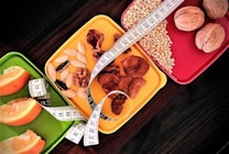 A selection of three colorful trays is filled with a variety of healthy snacks, including orange slices, nuts, seeds, and a measuring tape is artfully draped across them, suggesting themes of dieting or nutrition.