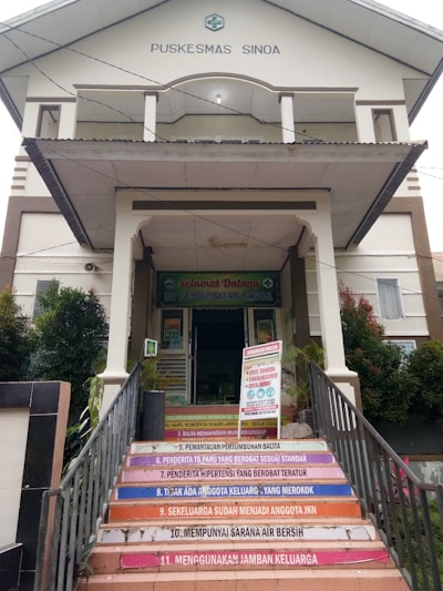 The image depicts a health care facility entrance labeled 'Puskesmas Sinoa'. The building has a welcoming sign and a set of colorful stairs leading up to it, each step containing a health-related message in Indonesian. Plants are seen on either side of the entrance.