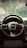 A detailed view of a car's steering wheel with the manufacturer's logo prominently centered. The dashboard and part of the interior are visible, with a background view through the windshield showing trees and a fence.
