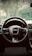 A detailed view of a car's steering wheel with the manufacturer's logo prominently centered. The dashboard and part of the interior are visible, with a background view through the windshield showing trees and a fence.