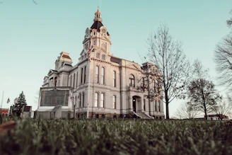 A grand historic courthouse with a clock tower and intricate architectural details stands surrounded by leafless trees. An American flag is prominently displayed near the entrance, and small flags decorate the surrounding monument. The sky is a clear blue, and the grass in the foreground is slightly frosted, suggesting a chilly season.