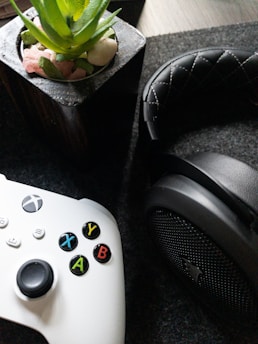 A white gaming controller with multicolored buttons is placed beside a black, cushioned headphone. In the background, there is a potted plant with green leaves and small stones in a container.