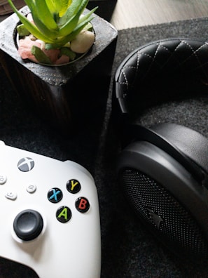 A white gaming controller with multicolored buttons is placed beside a black, cushioned headphone. In the background, there is a potted plant with green leaves and small stones in a container.