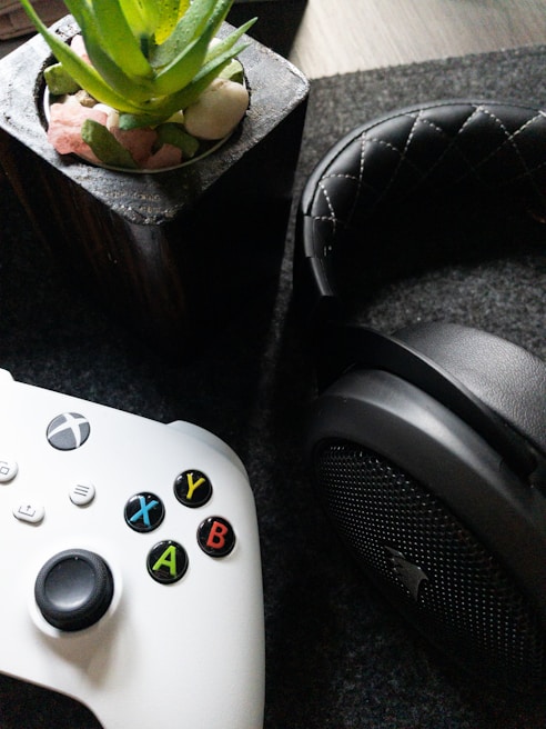A white gaming controller with multicolored buttons is placed beside a black, cushioned headphone. In the background, there is a potted plant with green leaves and small stones in a container.