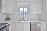 A modern and bright kitchen with white cabinetry and gold handles. A large window above the sink allows natural light to flood the room. Stainless steel appliances, including a dishwasher and an oven, are present. Minimalist decor and a small round sign that reads 'Kiss the Cook!' add a homey touch.