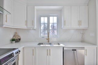 A modern and bright kitchen with white cabinetry and gold handles. A large window above the sink allows natural light to flood the room. Stainless steel appliances, including a dishwasher and an oven, are present. Minimalist decor and a small round sign that reads 'Kiss the Cook!' add a homey touch.