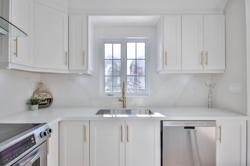 A modern and bright kitchen with white cabinetry and gold handles. A large window above the sink allows natural light to flood the room. Stainless steel appliances, including a dishwasher and an oven, are present. Minimalist decor and a small round sign that reads 'Kiss the Cook!' add a homey touch.