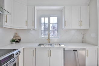 A modern and bright kitchen with white cabinetry and gold handles. A large window above the sink allows natural light to flood the room. Stainless steel appliances, including a dishwasher and an oven, are present. Minimalist decor and a small round sign that reads 'Kiss the Cook!' add a homey touch.