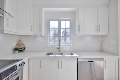 A modern and bright kitchen with white cabinetry and gold handles. A large window above the sink allows natural light to flood the room. Stainless steel appliances, including a dishwasher and an oven, are present. Minimalist decor and a small round sign that reads 'Kiss the Cook!' add a homey touch.