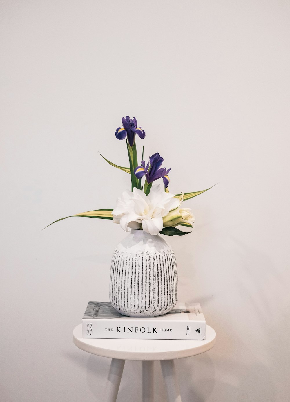 a white vase with purple and white flowers on top of a stack of books