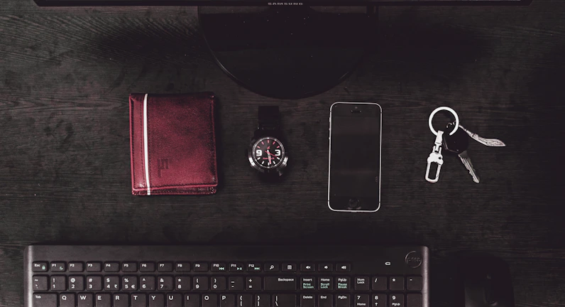 A sleek wallet, keys, and watch arranged neatly on a dark wooden desk with soft natural light.