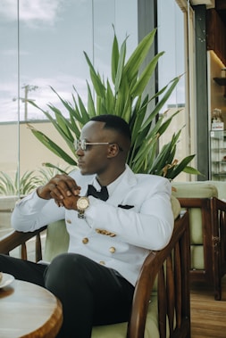 A man in a white blazer with gold buttons is seated in a wooden chair, wearing sunglasses and a wristwatch. He appears relaxed, resting his arms on the chair's armrests. The background includes a large green plant, glass windows, and indoor plants outside the window.