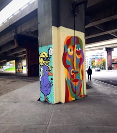 A vibrant urban street art piece on the pillar of an underpass, featuring a colorful abstract face in shades of purple, orange, green, and red. To the left, there is a cartoonish purple bird with a yellow beak and a humanoid figure with a thought bubble. The environment includes a concrete sidewalk and nearby vehicles and buildings.