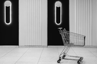 A shopping cart is placed on a tiled floor in front of a modern wall with vertical lines and glowing circular lights resembling exclamation marks. The overall setting is minimalist with a monochrome color scheme.