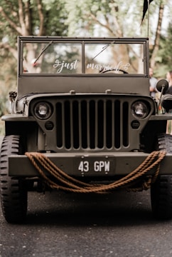 A vintage military jeep is adorned with a 'just married' sign on the windshield. The vehicle is parked on a paved path, surrounded by greenery. Thick ropes are coiled around the front, and the scene is bathed in soft, natural light.