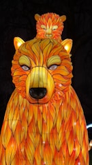 A brightly illuminated lantern displays a stylized bear with an exaggerated fur pattern in vibrant orange and yellow. A smaller bear is perched on top of the larger bear's head. The scene is set against a dark background, enhancing the glowing effect.