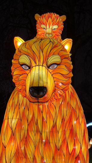 A brightly illuminated lantern displays a stylized bear with an exaggerated fur pattern in vibrant orange and yellow. A smaller bear is perched on top of the larger bear's head. The scene is set against a dark background, enhancing the glowing effect.