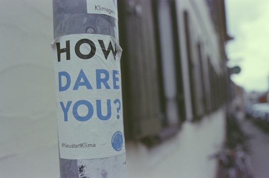 A poster with the text 'HOW DARE YOU?' is wrapped around a pole. The background features a blurred urban street scene with buildings and bicycles.