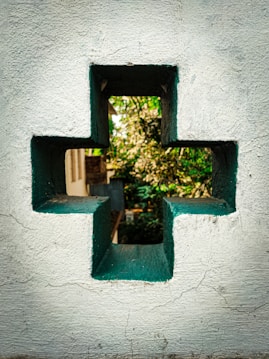 A cross-shaped cutout in a textured, white concrete wall. Through the opening, a glimpse of a garden with lush green foliage and a building can be seen in the background. The edges of the cutout are painted green, contrasting with the off-white wall.