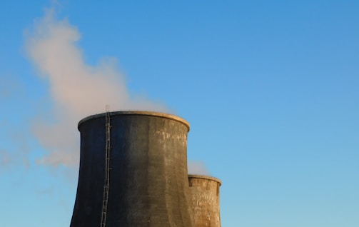 Two large cooling towers with steam rising from them are set against a clear blue sky.