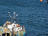 A small fishing boat docked at a pier, equipped with various antennas and fishing nets. The surrounding water is a deep blue, and a bright orange buoy floats nearby.