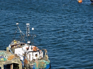 A small fishing boat docked at a pier, equipped with various antennas and fishing nets. The surrounding water is a deep blue, and a bright orange buoy floats nearby.