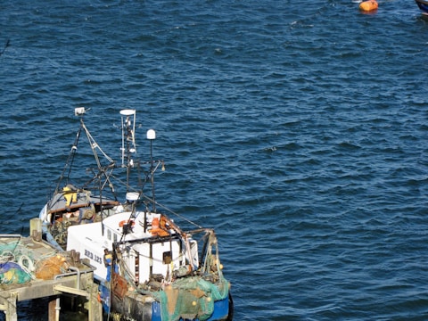 A small fishing boat docked at a pier, equipped with various antennas and fishing nets. The surrounding water is a deep blue, and a bright orange buoy floats nearby.